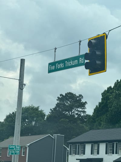 The image shows an intersection in Mountain Park, United States of America, marked by street signs and a traffic light. One street sign reads "Five Forks Trickum Rd". Another sign below it shows "Garner Road SW 1200" and "Five Forks Trickum Road SW 5330". The sky is overcast, suggesting cloudy weather. Trees and rooftops of buildings are also visible in the background.