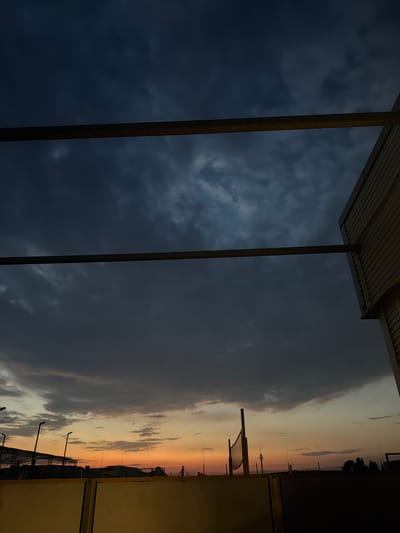 The image depicts a twilight scene, possibly taken from a balcony or elevated viewpoint, looking towards the horizon. The sky is filled with dark, brooding clouds in the upper portion, transitioning to lighter, more diffused colors of orange and yellow near the horizon, indicating either sunrise or sunset. In the foreground, two horizontal beams run across the frame, likely part of an overhead structure. To the right, a portion of a building with corrugated siding is visible. Below the sky, silhouettes of various objects can be seen, including what appears to be a volleyball net and poles, along with some distant structures and trees. The overall impression is one of a calm, atmospheric evening or early morning. There are no people visible in the image. The location is specified as Singen (Hohentwiel), Germany.