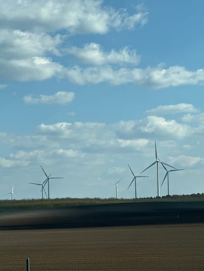 The image shows a rural landscape under a sky with scattered cumulus clouds. Several wind turbines are positioned along the edge of a cultivated field, located near the city of Ács, Hungary. The field has furrows and appears recently tilled. The lighting suggests it is daytime, with relatively clear visibility. The image has been captured from a slight angle or possibly rotated, making it appear as though the landscape is tilted to the right.