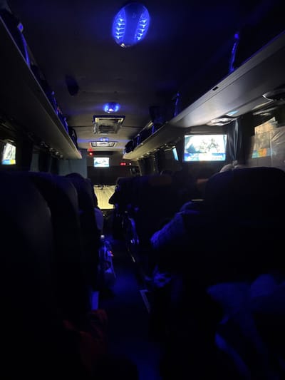 The image depicts the interior of a passenger bus or coach under dim, predominantly blue ambient lighting. Numerous unidentifiable individuals are seated throughout the vehicle, appearing as dark silhouettes in the seats, facing forward.
**Environment:** The bus features a central aisle separating two rows of passenger seats. Overhead luggage compartments line both sides of the interior. The ceiling is dark, punctuated by several circular blue LED light fixtures that cast a strong blue glow onto the ceiling and parts of the aisle. Several small flat-screen monitors are mounted within the cabin; one prominent screen on the right displays what appears to be a broadcast or media content featuring a person's head and visible text. Another screen is visible on the left, and two smaller screens are further forward near the front of the bus. A red digital display, located above the central aisle towards the front, shows "22:12". Curtains or window coverings are drawn along the sides of the bus.
**People:** Passengers occupy many of the seats, their specific features obscured by the low light. They are stationary, presumably traveling. One passenger in the mid-ground on the right side appears to be wearing a light-colored or patterned garment on their upper body. A red object, possibly a bag or blanket, is visible on the floor near a passenger in the foreground on the left.
**Objects:** The seats are dark-colored. The blue ceiling lights have a distinctive internal finned or grid-like pattern. The floor of the aisle is dark, with subtle reflections of the blue light.
**Actions/Interactions:** The passengers are seated, but no specific actions or direct interactions are discernible due to the darkness and distance. Some individuals appear to be passively observing the screens.
**Location:** The image provides no visual information to confirm the specific geographical location within the city of Sedanovo, Russia. The scene is identifiable only as the interior of a bus.