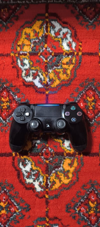 A black PlayStation 4 controller rests on a red, intricately patterned rug. The rug features a repeating design in yellow, white, and dark gray, creating a visually complex background. The controller is centrally positioned, with its directional buttons and analog sticks clearly visible. A thin blue light bar is illuminated on the top of the controller. The overall scene is static and devoid of human presence. This image is located in Urgench, Uzbekistan.