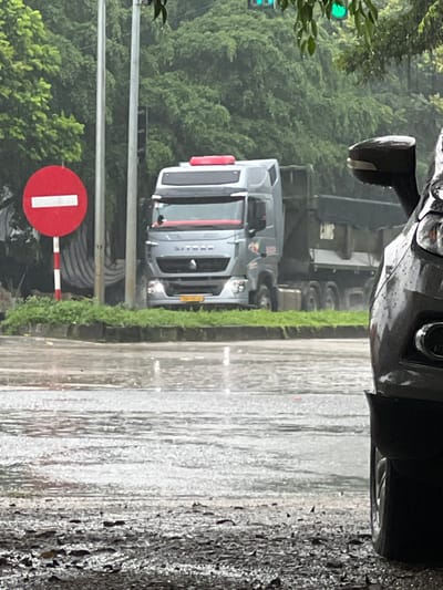 The image captures a street scene in Tam Dương, Bắc Ninh, Vietnam, during what appears to be heavy rainfall. A grey semi-trailer truck, identified by the "SITRAK" logo on its grille, is prominently featured in the mid-ground. The truck has a red light bar on its roof. To the left of the truck, a circular red traffic sign with a horizontal white bar indicates "Do Not Enter". A dark-colored car is partially visible on the right side of the frame, its front end showing a headlight and part of the grille. The road is wet and reflects the overcast sky, with visible puddles and a glistening surface. Dense green trees form a backdrop, suggesting a lush, possibly tropical environment. The traffic signal above the truck shows a green light. The overall atmosphere is damp and somber due to the persistent rain.