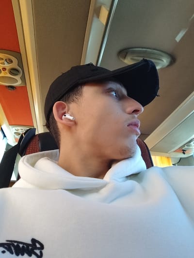 A young man wearing a black baseball cap and white hoodie sits on a bus. He has fair skin, a light beard, and an AirPod in his left ear. He is looking to his right with a neutral expression. The interior of the bus is visible in the background, including the overhead compartments and ceiling lights. The bus seat next to him appears to be empty.
