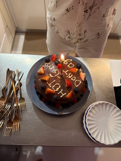 A chocolate cake decorated with white frosting, strawberries, blueberries, and raspberries sits on a blue plate. The white frosting spells out "HOT OG 100". A single lit candle sits in the center of the cake. A stack of forks is to the left of the cake, and two scalloped plates are to the right. The cake is on a metallic surface, possibly a countertop, with a blurred background showing a kitchen area and a person wearing a white shirt with a floral pattern and white shorts. The scene appears to be indoors. There is no information in the image to determine its location in Chang Phueak, Thailand.