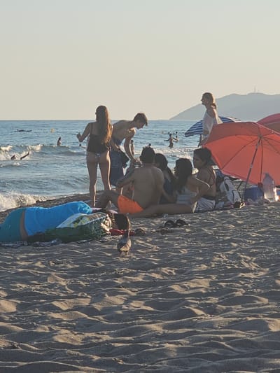 The image depicts a group of people on a sandy beach, with the sea in the background. The photograph is oriented vertically. Several individuals are visible, some standing, others sitting or bending down. One person in the foreground is wearing a blue garment. There is an orange umbrella providing shade for some of the people. The sand has a textured appearance with ripples.

The scene appears to be during the daytime, with bright sunlight casting shadows. The sea looks calm. The presence of beachgoers, umbrellas, and the ocean strongly suggests a recreational setting at a beach.

One person in the background is wearing a white top with thin straps. Another person is wearing orange shorts. A few small birds are visible in the water. The overall mood seems relaxed and social, typical of a day spent at the beach.

There is no visible text in the image. The location context suggests this is likely Castelldefels, Spain, a known beach destination.