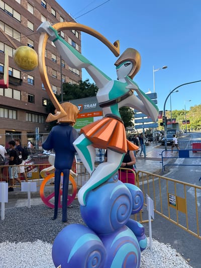 This is a medium shot, outdoors, low-angle photo taken in what appears to be a city street. The main subjects are two large, stylized figures that are part of a street art installation. The figure in the foreground is a woman with a golden helmet, wearing a teal and white outfit and an orange skirt. She is holding a large golden bow and arrow, with a yellow sphere suspended below it. Behind her, a second, smaller figure is dressed in a dark blue outfit. Both figures are made of brightly colored, geometric shapes.
The figures are set against a backdrop of a multi-story brick building on the left and a street with cars, people, and barriers on the right. The sky is a clear, bright blue, suggesting it is a sunny day. The time of day appears to be late morning or early afternoon. The scene is likely part of a festival or public art display, given the scale and elaborate nature of the figures and the presence of temporary barriers.
Visible text includes "dorsia," "frenda," "nink," "utiful," and a logo for "T TRAM METROPOLITA D'ALACA" with "FGV" and "TRAM" brands. There are also phone numbers and advertising-like text in Spanish. The location context of Alicante/Alacant, Spain, is given. The overall mood is vibrant and festive, with the bright colors and dynamic poses of the figures.