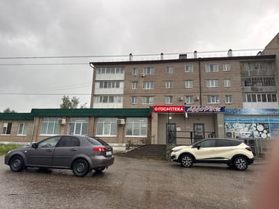 This media file captures an outdoor urban scene featuring two cars parked on a wet asphalt surface in front of a multi-story brick building under an overcast sky. The overall impression is one of a cloudy, possibly rainy day.

The primary subject is a four-story brick apartment building with numerous windows, some of which feature white frames and external air conditioning units. The upper part of the building has multiple balconies, some enclosed, others open and appearing somewhat dilapidated. Below the main building, a single-story structure with a dark green sloped roof extends outwards. This lower building also features large windows and air conditioning units.

Two cars are prominently visible: a dark gray sedan (appears to be a Chevrolet Lacetti) parked on the left, facing away from the viewer, and a cream-colored SUV with a black roof (likely a Renault Kaptur) parked on the right, facing left. Both vehicles are on a visibly wet ground, suggesting recent rain. Utility lines are stretched across the upper portion of the frame.

Several signs are affixed to the buildings. A prominent red sign reads "ГОСАПТЕКА" (GOSAPTEKA), indicating a state pharmacy. Next to it, a blue sign displays "АССОРТИ" (ASSORTI), likely a shop name. Further right, another blue sign reads "ПРОДМАГИЯ" (PRODMAGIYA), which translates to "Food Magic," indicating a grocery store. Below "ПРОДМАГИЯ," smaller text is visible: "ТОРГОВАЯ СЕТЬ" (TORGOVAYA SET' - Trading Network), "Товары, доступные каждому" (Goods available to everyone), "Домашняя выпечка" (Homemade pastries), and "Продукты местных производителей" (Products from local manufacturers).

No people are visible in the image, and there is no discernible activity taking place. The time of day appears to be daytime, given the ambient light, though obscured by cloud cover. The Russian text on the signs confirms the location context of Mozhga, Russia.