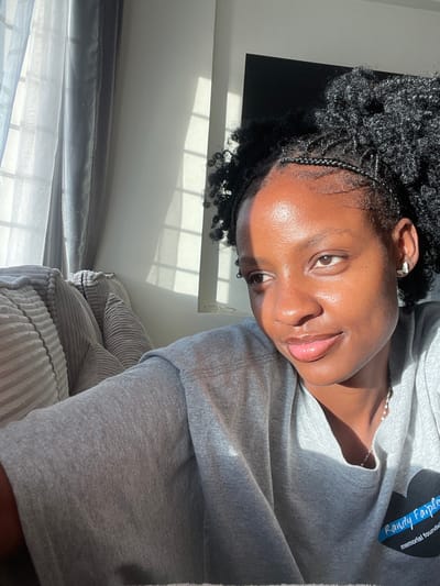 The image depicts an indoor portrait of a female individual. She appears to be of African descent, possesses dark skin, and has black curly hair styled with small braids along the hairline, pulled back into a ponytail or bun. Her facial expression is neutral, with her gaze directed towards the upper right of the frame. She wears a grey short-sleeved t-shirt, a silver-toned necklace with small, reflective elements, and a light-colored earring, possibly butterfly-shaped, in her visible right ear. On the left side of her t-shirt (viewer's right), a blue heart-shaped logo with white text reading "Randy Faiple memorial foundation" is present.

The environment appears to be a room with light-colored walls. Strong natural light, originating from a window on the left, illuminates the scene, casting distinct shadows of window panes onto the wall and curtains. The window features both sheer light-colored curtains and darker drapes. A portion of a light-colored, ribbed, upholstered sofa or chair is visible on the far left. In the background, behind the individual's head, a dark, rectangular object, possibly a television screen or furniture, is partially visible. The image provides no visual indicators to confirm the specific location within Thika, Kenya.