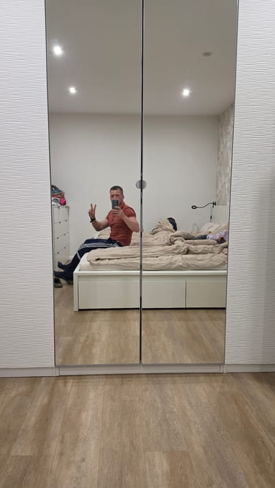 The image shows a reflection in a mirrored closet. A man is taking a selfie in the mirror. He is wearing a reddish-brown t-shirt and black track pants with white stripes. He is making a peace sign with his left hand. The reflection also shows part of a bed with a beige comforter, a white dresser with clothes on top, and a light-colored wooden floor. The top and bottom of the closet doors have a textured white panel. The location is a bedroom in Ihringshausen, Germany.