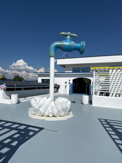 The image depicts a rooftop installation in Baliovtsi, Bulgaria, featuring a large, blue faucet sculpture supported by a base of white, cupped hands. The sculpture is positioned on a gray, tiled surface. In the background, a white building with blue trim and accents is visible, along with a section of white latticework with a yellow railing. The sky is clear blue with scattered white clouds. The scene appears to be outdoors, under bright daylight.