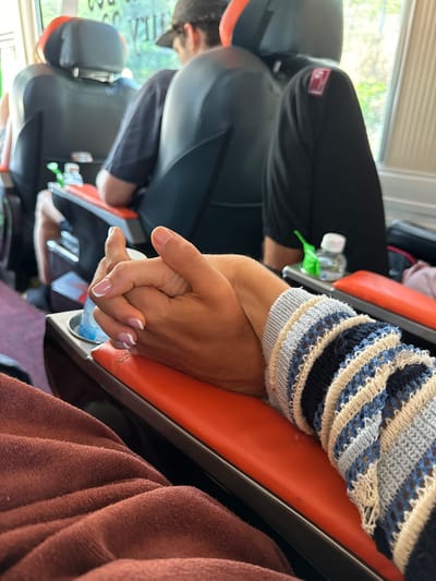 The image captures an intimate moment inside a passenger vehicle, likely a bus or van, during daytime travel in Phan Son, Vietnam. In the foreground, two hands are clasped together, symbolizing affection or comfort between two individuals. One arm, partially visible, has a darker skin tone and is covered by a patterned knit sleeve featuring blue, white, cream, and black stripes. The other hand, with a lighter skin tone and neatly manicured nails with white French tips, is held gently.
The setting is an interior space dominated by orange and black seats, typical of passenger transport. Several other passengers are visible in the midground and background, sitting in similar seats, though their faces are largely obscured or blurred. One person in the background wears a black cap. Small plastic water bottles are visible tucked between seats. A brown or maroon textured fabric, possibly a blanket or clothing, occupies the far right of the frame.
Bright natural light streams in from a large window on the left side, indicating daytime. Outside the window, a blurred view of lush green foliage and a bright sky suggests an outdoor, possibly rural or scenic, environment. Window blinds are visible at the top of the window. A small, red rectangular label with unidentifiable white text or a logo is attached to the back of a seat headrest. The overall scene depicts travel and companionship.