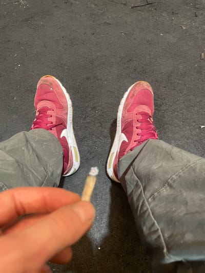 The image shows a person's perspective from a seated position, focusing on their feet and hands. The person is wearing dirty, gray pants and red sneakers. In their right hand, they are holding a lit cigarette with a glowing ember. The background appears to be a dark, gritty surface, possibly asphalt or concrete, with some scattered debris. The scene suggests an outdoor urban environment, but no specific landmarks or details are visible to pinpoint a location within Gerstheim, France.