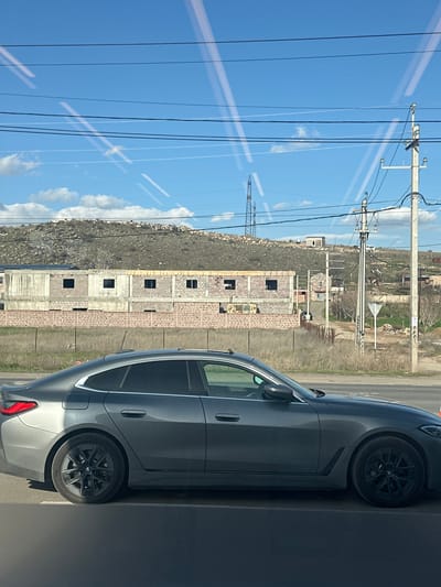 A medium-level shot depicts a view from inside a vehicle, showing the reflection of electrical wires and a bright, partly cloudy sky on the left side. The reflection is overlaid on an external view that includes a partially constructed building made of light gray blocks, possibly in a rural or developing area of Zovuni, Armenia. A rocky hill sits adjacent to the construction site. A gray car is positioned on the right, with its wheels and side visible, suggesting the photo was taken from inside a moving vehicle. The overall environment appears to be a mix of natural landscape and urban development.