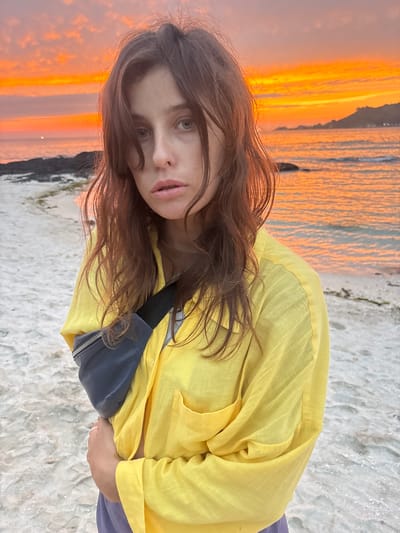 This is a photograph of a woman standing on a sandy beach during sunset. The woman has long, brown hair and is looking directly at the camera with a neutral expression. She is wearing a bright yellow, long-sleeved shirt and a dark blue fanny pack across her chest. Her lower body is mostly out of frame, but she appears to be wearing purple pants. The background is dominated by the ocean and the sky, which are illuminated by the warm, vibrant colors of the sunset. The sky is a gradient of orange and pink, with wispy clouds. The ocean reflects the sunset's colors, creating a shimmering effect. The beach sand is light in color and appears textured. The overall mood of the image is serene and peaceful, capturing the beauty of a sunset on a beach. The image appears to be taken in Jeju, South Korea, given the provided location context. There is no visible text in the image.
