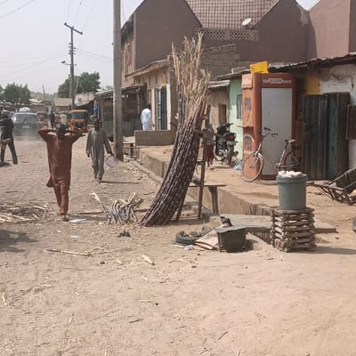 The scene depicts a daytime street view in Kaura Goje, Nigeria, characterized by an unpaved, dusty thoroughfare. In the mid-ground, a large bundle of sugarcane stalks leans against a wooden stand on the right side of the dirt road, with scattered smaller pieces of sugarcane and debris on the ground directly in front. A grey bucket, containing white ovoid objects, rests on a stack of wooden planks on the far right. Adjacent to this, a light-colored bicycle is parked next to an orange-brown "POS" (Point of Sale) kiosk, which stands against a light green building. A dark motorcycle is parked further back on the right.
On the left side of the dirt road, two men are visible. The man in the foreground wears a brown garment and has his hands placed on or near his head while walking towards the viewer. Behind him, another man in a light-colored garment walks away from the viewer, carrying a light-colored bowl in his left hand. In the background on the left, several vehicles, including a dark sedan and a yellow auto-rickshaw, are visible on the street. Utility poles with overhead wires are present on the left side of the road. Buildings with varying architectural styles, including a multi-story brown building with a tiled roof and single-story structures, line both sides of the street. A man in a white garment stands in a doorway of a building on the right side of the mid-ground. The sky is clear and light.
