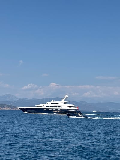 An extremely large, dark blue and white motor yacht is cruising through choppy blue water under a clear sky with scattered clouds. The yacht is positioned in the right-middle of the frame, moving from left to right. It has multiple decks and several windows. A smaller, dark tender boat is following behind the larger yacht, creating a wake. In the background, a hazy, mountainous coastline is visible, with two smaller white boats in the distance. The overall scene appears to be set in a coastal area of Turkey.
