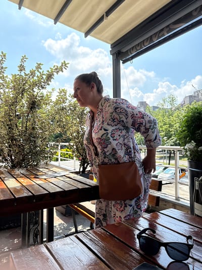 The image depicts an outdoor seating area of a commercial establishment located in Rakovica Urban Municipality, Serbia, under a bright, partly cloudy sky. One female individual is visible in a profile view, oriented towards the left. She has dark hair styled in an updo and is wearing a long-sleeved, collared dress with a white base and a multi-colored floral and paisley pattern in shades of blue, pink, and green. A brown leather cross-body bag is slung over her right shoulder, resting at her hip, and her right hand is partially gripping the fabric of her dress near her hip.
The foreground features two dark-stained wooden slatted tables. The table on the right has a pair of black sunglasses resting on its surface. The table on the left contains a clear glass ashtray. The surfaces of both tables appear glossy, reflecting sunlight. An overhead structure, comprising a light beige fabric awning supported by dark gray beams, extends across the top of the frame. Below the awning, a dark roller blind with a light-colored, indecipherable pattern is partially visible.
To the left of the woman, several bushes or small trees with light green leaves are planted. To her right, a dark potted plant with small white flowers sits on the table, and a black bucket or container, partially revealing the text "PROSECCO," is visible on the ground. A white metal railing separates the seating area from a lower level, revealing a street with parked cars, including a prominent white vehicle, and distant urban buildings against the blue sky with scattered white clouds.