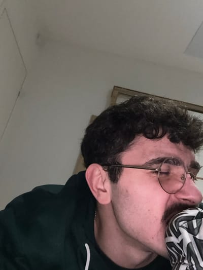 A man with curly dark hair and glasses is angled towards the right side of the frame. A patterned cloth or fabric covers part of his face. He wears a dark green hooded sweatshirt with a white design on the sleeve. The background is a plain white wall. A partially visible window frame can be seen behind the man’s head. The lighting in the room is soft and diffused.