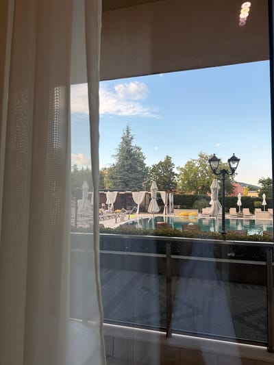 The image shows an outdoor swimming pool area seen through a window, with sheer white curtains in the foreground partially obscuring the view. The scene is set in Hisarya, Bulgaria, and appears to be during the daytime, with clear blue skies and some scattered clouds. Sunlight is casting shadows, suggesting it's not midday.
The pool itself is a prominent feature, with blue water and lounge chairs around it, some of which are covered by white umbrellas. There are also decorative lamp posts and lush green trees bordering the pool area. In the background, buildings and hills are visible, hinting at a resort or hotel setting. The reflection on the window adds another layer to the scene, showing a distorted view of the sky and greenery. There are no people visible in the image.
The overall mood is tranquil and inviting, suggesting a pleasant day for relaxation by the pool. The presence of the curtains and the way the light filters through them creates a soft, slightly diffused atmosphere. There is no visible text in the image.