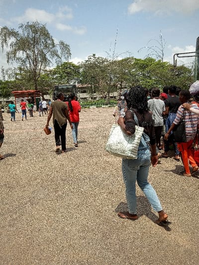 The image shows a group of people in a paved area. They are standing and walking near a bus. One person is wearing a red shirt and walking toward the left side of the image. Another person is carrying a large white bag and walking toward the right side of the image. The image is taken from a slightly elevated perspective. The location of the image, as determined by the coordinates, is Ota, Nigeria. There are trees on the periphery and a single-story building in the background. There is a bus stop with a red roof in the left foreground.