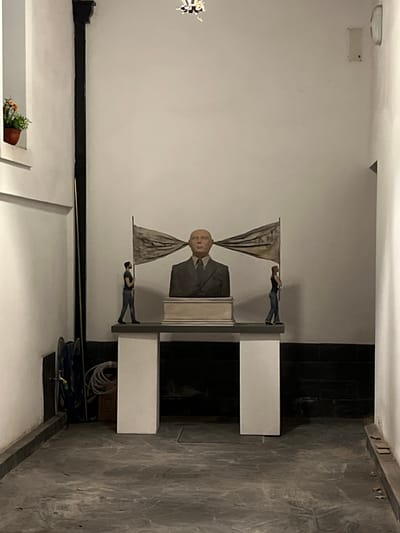 The image depicts a sculpture of a bald man in a suit, with draped fabrics resembling flags behind him. Two miniature figures are positioned atop the flags, as if carrying them. The sculpture is mounted on a white pedestal. The background consists of a white wall, a dark-tiled area, and a section of patterned grey flooring. A plant in a pot is visible in the lower left corner. A coil of blue and white cable sits beside the sculpture. The sculpture is located in Bahía Blanca, Argentina.