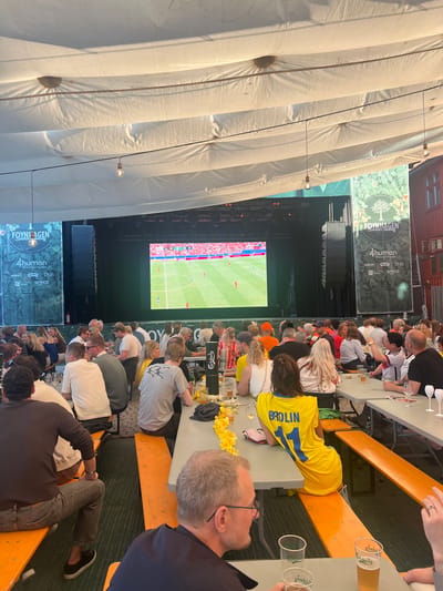 The image depicts a lively outdoor gathering, likely at a public screening event, under a large white canopy. A large screen at the back displays a football match, with a crowd of people seated at long tables, facing the screen. The setting appears to be the Foynhagen Summer Garden in Tønsberg, Norway, as indicated by signage on a banner to the left of the screen. The crowd consists of men and women of various ages, many dressed casually, some in football jerseys. They are engaged in watching the match, socializing, and enjoying drinks. The tables are set with glasses, bottles, and some are adorned with yellow flowers, suggesting a festive atmosphere. The lighting suggests it is daytime, possibly late afternoon or early evening. The overall impression is of a communal sporting event experience.