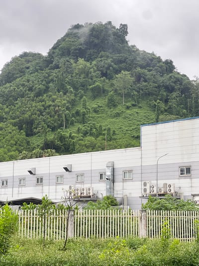 This image depicts a building with a lush, green mountainside in the background. The building appears to be a modern industrial or commercial facility with a light-colored facade and multiple windows. Several outdoor air conditioning units are visible on the exterior of the building. In front of the building is a white metal fence, behind which are various plants and vegetation. The mountainside is densely covered with trees and foliage, with some mist or clouds obscuring the upper reaches. The sky is overcast. The scene is situated in Sơn Dương Commune, Vietnam.