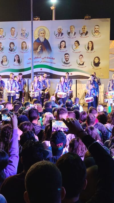 A crowd watches a drumming band perform on stage. Behind the band is a large banner featuring a central religious figure labeled "ST ELIAS THE PROPHET" and several portraits of individuals above and below him. Arabic text and the flag of Syria are also on the banner. A date, "22/06/2025," is visible, along with other smaller Arabic text and additional names. People in the audience are holding up phones to record the performance.