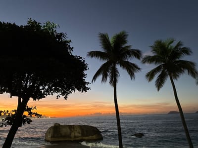 The image displays a coastal scene at dusk, characterized by a vibrant sunset gradient in the sky. The upper sky is a deep twilight blue, transitioning to orange and yellow hues at the horizon, where subtle dark clouds are present. In the immediate foreground, a large, dark tree with distinct foliage is silhouetted on the left. To the right, two tall palm trees with visible fronds are silhouetted against the sky. The midground features a dark blue ocean with visible waves breaking, showing white foam near the shore. A large, light-colored rock formation is situated in the water on the left, with waves washing around its base. On the distant horizon to the left, a cityscape is visible, marked by numerous illuminated buildings and the dark silhouettes of hills or mountains. To the right on the horizon, dark landmasses or islands are silhouetted. No individuals are visible in the scene. The distinct geographical features, including the mountains, city lights, and coastal setting at sunset, strongly indicate the location as Ipanema Beach in Rio de Janeiro, Brazil, likely looking towards the west (towards Leblon and Vidigal/Dois Irmãos mountains).