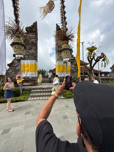 In the city of Baturiti, Indonesia, a person wearing a black cap and black shirt is seen from behind, holding up a smartphone to capture an image. The phone is pointed towards a traditional Balinese temple gate, characterized by its stone structure and intricate carvings. Two large white and yellow banners hang prominently on either side of the gate. Above the gate, decorative elements made of dried leaves and woven materials are suspended, along with a long yellow flag. In front of the gate, several people are visible. On the left, a woman in a denim skirt and a light-colored top stands on a paved area. Further back, a man and a woman in colorful traditional attire are walking up the temple steps. The ground in front of the temple is paved with large gray stone tiles. The sky is overcast with visible clouds. The overall scene captures a moment of cultural tourism at a significant architectural landmark.