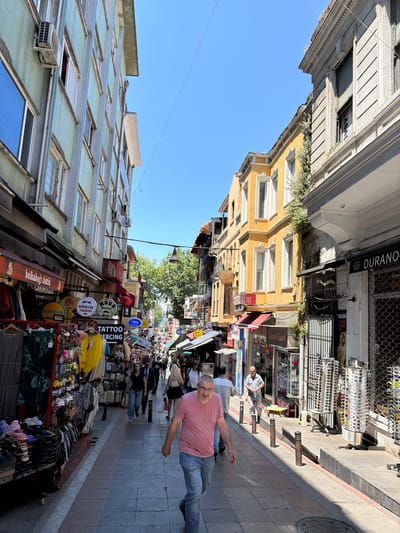 A street scene in Kadıköy, Turkey, likely during the daytime on a sunny day. The perspective is a downward-looking shot, with buildings on both sides of a pedestrian walkway. The buildings are a mix of styles, with some appearing older with ornate architectural details and others more modern. One building is painted bright yellow. Shops line the street, displaying various goods like clothing, sunglasses, and what appears to be souvenirs or trinkets. Signs are visible in Turkish, including "CANIKUGLO", "balkabağı butik", "IS MERKEZİ", "GÖZLÜK TAMİRİ", "MODA", "TATTOO ERCING", "ASAS", and "DURANO".
Several people are present on the street, some walking and others appearing to browse the shops. A man in a pink t-shirt and jeans is prominently featured in the foreground, walking towards the camera with an outstretched arm. Another man in a checkered shirt is also visible. The atmosphere seems to be that of a busy market or shopping district. The sun casts shadows on the ground, indicating it's not early morning or late evening.