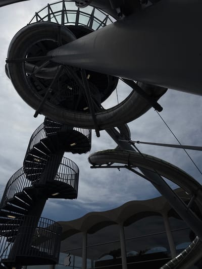 This is a low-angle shot of a large, modern outdoor art installation featuring a winding metal slide and spiral staircase in Aventura, Florida. The sculpture is predominantly gray metal with some translucent elements. The slide is a large, tubular structure that curves and twists, appearing to be part of a larger, complex design. A dark metal spiral staircase with railings is visible on the left side of the frame, ascending towards the slide structure. The sky above is cloudy, with patches of blue visible. In the background, the distinctive wavy roofline and large glass windows of a modern building, likely a retail space, are visible. An Apple logo is clearly visible on the glass facade of the building. The overall impression is one of contemporary architecture and public art.