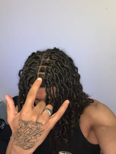 The image shows a person with dark, curly locs making a hand gesture with their right hand. They are wearing a ring on their ring finger and have a tattoo on the back of their hand. The person appears to be wearing a black tank top. The background is a plain, light-colored wall. There is no evidence to support that the image was taken in Massy, France.