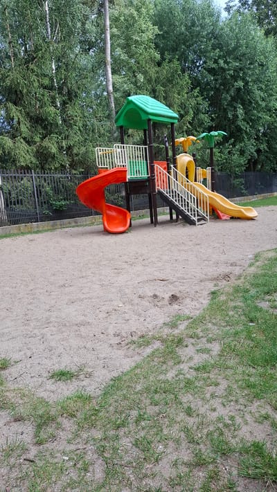 The image depicts a section of a playground with a multi-level play structure. The structure features bright plastic components in green, yellow, and orange, including slides and platforms. A yellow, wavy slide is visible at the top, and a red, curved slide is at the bottom. White stairs with black railings connect different levels of the play equipment. The playground is situated on a sandy ground with patches of green grass. In the background, a dense cluster of green trees and a dark metal fence are present. The lighting suggests it is daytime, likely with overcast conditions given the absence of strong shadows. The scene appears calm and unoccupied, implying no specific activity or event is taking place at the moment the photo was taken. There is no visible text in the image. The location context provided is Grójec, Poland.