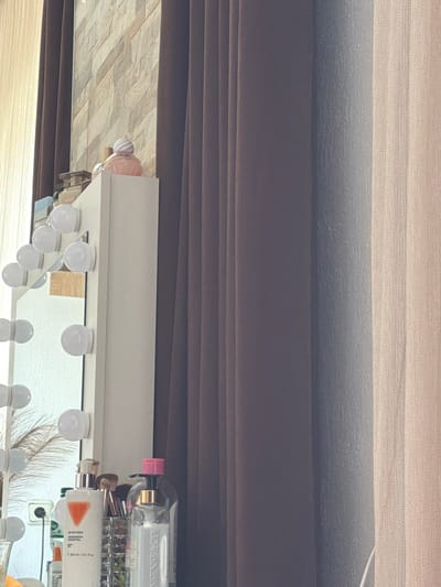 The image shows a vanity mirror with lights, a wall decorated with stacked beige and brown rectangular tiles, and several items on a white surface below the mirror. Among these items are bottles of what appear to be skincare or perfume products, makeup brushes, and a decorative perfume bottle with a pink swirl top. The vanity is positioned next to dark brown curtains. To the right of the brown curtains, there are sheer, light beige curtains. The scene is indoors, likely in a personal space such as a bedroom or dressing room. There are no people visible in the image. The location in Zmeitsa, Bulgaria, is not evident from the visual information provided in the image itself.