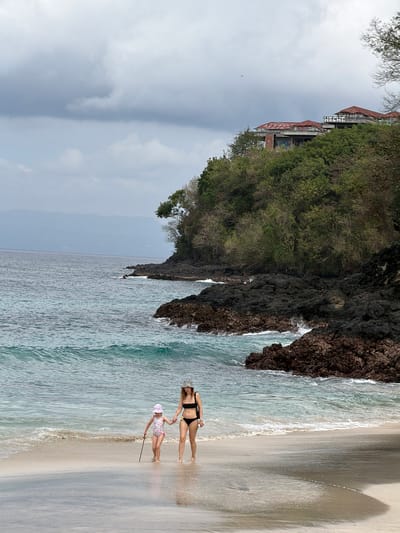 Two people, an adult and a child, are on a sandy beach next to the ocean. The adult is wearing a black bikini and a leopard print hat, and is lying on their back. The child is wearing a pink and white swimsuit and a matching hat, and is standing up holding a stick. The ocean water is light blue and there are small waves. The beach is light brown sand and there is a rocky cliff on the left side of the image with green trees growing on it. In the background, there is a building with a red roof. The sky is overcast and cloudy. The image appears to be taken in the daytime. There is no visible text in the image.