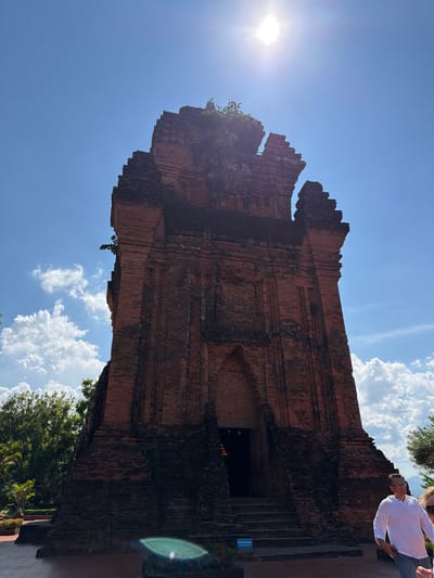 In the city of Tuy Hoa, Vietnam, an ancient brick tower stands against a bright blue sky with scattered white clouds. The sun is high in the sky, casting a bright glare in the upper right portion of the image. The tower, constructed of weathered red bricks, has a tiered, stepped structure and appears to be of Cham origin, indicated by its architectural style. Vegetation is visible on the upper parts of the tower and in the surrounding area. In the foreground, a person wearing a white long-sleeved shirt and jeans walks away from the camera, with the top of another person's head visible to their right. A blue sign with white lettering is positioned at the base of the tower's entrance stairs. The ground in front of the tower is paved with reddish-brown tiles.