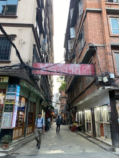 The image depicts a narrow, cobblestone street in Kathmandu Metropolitan City, Nepal, flanked by multi-story buildings on both sides. The architecture on the left features a beige facade with dark brown trim and barred windows, while the building on the right is constructed of red brick with numerous windows, some of which are open. A large, weathered red sign with white text is suspended across the street between the two buildings. Numerous electrical wires and conduits are strung haphazardly between the buildings, contributing to a cluttered overhead view.
On the left side of the street, businesses are visible, including one with a sign indicating "THAMEL HIMALAYAN GUEST HOUSE" and "Precious Care Collection." Displays of jewelry and currency exchange rates are present. A man in a white shirt and light-colored pants, carrying a blue backpack, walks away from the camera down the street.
On the right side of the street, shops with illuminated storefronts display clothing on racks. A person in a green shirt is seated on the curb in front of one of these shops. Further down the street, two individuals are walking away from the camera, and other figures are visible in the distance. The overall scene suggests a bustling urban environment, typical of commercial areas in Kathmandu. The presence of air conditioning units mounted on the buildings and the style of the signage further place the location within the city.