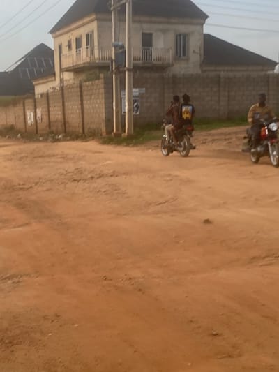The image depicts an unpaved, reddish-brown dirt road in Iddo Tudunwada, Nigeria. The foreground consists of the uneven earthen surface, displaying variations in texture and minor ruts. In the mid-ground, two motorcycles are in motion. To the left, two male individuals are seated on a dark motorcycle, traveling away from the viewer; the passenger wears a dark shirt with a yellow graphic on the back. To the right, a single male individual on a red and black motorcycle is partially visible, moving towards the right side of the frame; he wears a light-colored shirt.

The background features a two-story building with a light-colored facade, dark sloped roof, and a white-railed balcony on the upper floor. A concrete block wall, topped with security wire, runs horizontally across the mid-ground behind the motorcycles, defining the boundary of the property where the building is located. Utility poles with overhead lines are visible along the wall. Minimal, dry-looking vegetation is present at the base of the wall. The sky is indistinct, suggesting diffused daylight.