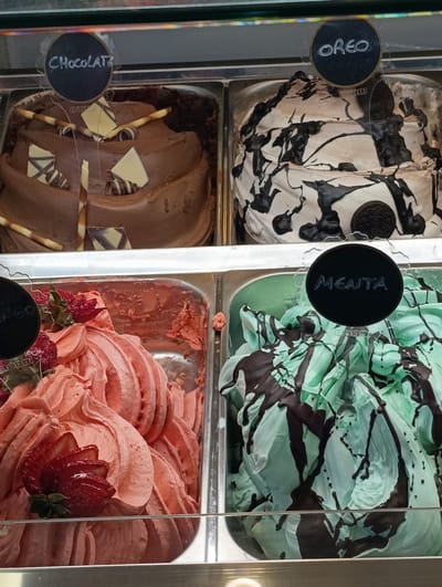 This media file shows a close-up, eye-level view of four large metal trays filled with gelato, displayed in a refrigerated case. The gelato is presented in a variety of flavors and textures.
The top left tray contains chocolate gelato, garnished with chocolate sticks, white chocolate squares, and swirled chocolate wafers. A black circular sign above it reads "ChocolaTe?".
The top right tray holds what appears to be cookies and cream or Oreo gelato. It is a white gelato base with dark chocolate swirls and chunks, and whole Oreo cookies are visible. A black circular sign above it reads "OREO".
The bottom left tray is filled with strawberry gelato, topped with sliced fresh strawberries and some green leaves. A black circular sign is partially visible, with some text obscured, but the visible letters suggest "LANGO".
The bottom right tray contains mint chocolate chip gelato, characterized by its light green color and dark chocolate drizzles. A black circular sign above it reads "MENTA".
The overall scene suggests a gelato shop or an ice cream parlor. The lighting appears to be artificial and bright, typical of indoor display cases. The focus is solely on the gelato, with no people visible. The setting is clean and well-maintained, highlighting the appealing presentation of the desserts. The visible text indicates different gelato flavors offered. The context provided places the location in Odivelas, Portugal.