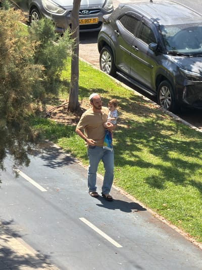 The image shows a man holding a baby while walking on a sidewalk next to a road. They are on a sunny day, with trees and grass surrounding them. Two cars are parked on the side of the road. The man is wearing a light brown t-shirt, blue jeans, and sandals. He also has a white kippah on his head. The baby is wearing a blue and white striped shirt. The license plate of the car in the foreground is partially visible and appears to be from Israel, with the numbers "45.967.11". The overall scene suggests a casual stroll in an urban environment.