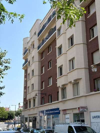 This is a street-level photograph, taken from a low angle looking upwards, of a multi-story building. The building has a facade of alternating sections of reddish-brown brick and cream-colored stucco. The stucco sections feature projecting horizontal bands of varying depths, creating a layered effect. There are numerous windows visible across the facade, some with dark frames and others appearing to be recessed. Small balconies with dark railings are present on some levels. In the foreground, on the left side of the image, is a cluster of green leafy branches, suggesting that the photograph was taken during a time of year when trees are in full foliage. The sky above is a clear, bright blue, indicating daytime.

On the ground floor of the building, there are several commercial establishments. One prominent sign reads "LAVERIE LIBRE-SERVICE" in large letters, indicating a self-service laundry. Other signs are partially visible, including "MACHINES A LAVER" (washing machines) and what appears to be a "Point d'Info Stationner à Courbevoie" (Information point Parking in Courbevoie), suggesting the location is Courbevoie, France, and possibly related to parking information or services. A white commercial van is parked on the right side of the street, partially obscuring the ground-level storefronts. Streetlights and traffic signs are visible further down the street, hinting at an urban environment. The overall impression is a mundane urban street scene on a bright, clear day.