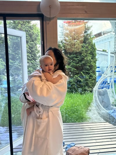 A person in a white robe is holding a baby, likely indoors. Outside, a garden is visible through a window, including grass, trees, and a white wicker chair. Based on the location Чёрное, Russia, and the reflection in the window, the building with red accents and lights could be a restaurant or establishment named "The Black Pearl."