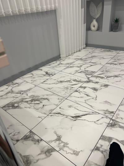 The image depicts a modern interior space with a floor tiled in a white marble pattern. The room appears to be an indoor environment, likely a living area or hallway. The walls are painted a neutral grey. To the right, there is a recessed shelf displaying a white decorative vase with a unique, circular opening and a silvery, leaf-like ornament. Next to it, another shelf holds a small potted plant in a clear glass container. White vertical blinds hang along the left side of the frame. The bottom right corner of the image shows a dark shadow cast by a person's foot, suggesting the presence of an individual in the scene. The lighting is even, indicating an indoor setting with artificial or diffused natural light. The overall impression is one of clean, minimalist decor. The location in Novi Iskar, Bulgaria, is not visually discernible from the image itself, as it is an interior scene lacking any external markers.