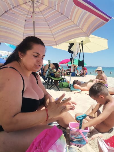 This image shows a sunny day at the beach in Kranevo, Bulgaria. The scene is filled with people enjoying the warm weather and the sea. In the foreground, a woman in a black bikini is sitting on the sand, applying sunscreen to her arm. Next to her, a young boy is playing with a pink beach bucket and spade.

Behind them, several beach umbrellas provide shade. Under one of the umbrellas, a man is lying down, possibly resting or sunbathing. Further back, a woman wearing a straw hat and bikini is sitting, and a man in a red shirt is kneeling. Other beachgoers are visible in the background, some swimming in the ocean, while others are relaxing on the sand.

The sky is clear and blue, indicating good weather. The presence of beach umbrellas, buckets, and people in swimwear clearly places the scene at a popular tourist destination during the summer. The overall atmosphere appears relaxed and recreational. No visible text is present in the image.