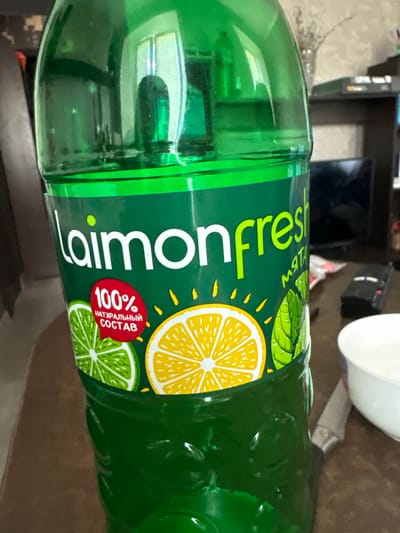 A close-up, eye-level shot features a large, green plastic bottle of "Laimon fresh" beverage. The bottle is translucent, allowing the green liquid inside to be visible. The label on the bottle is predominantly green with white and yellow lettering. Prominently displayed is the brand name "Laimon fresh." Below this, the text "100%" is enclosed in a red oval, with smaller Russian text that translates to "natural composition" beneath it. To the left of the red oval is an illustration of a sliced lime, and to the right is an illustration of a sliced lemon and mint leaves. The bottle appears to be on a brown wooden surface, possibly a table. In the background, a dark shelf with electronic equipment and other indistinct objects is visible. To the right of the bottle, a white bowl and the edge of a knife are also present on the surface. The lighting suggests an indoor environment, possibly a home or store. There is no indication of the specific location within the city of Kuvshinovo, Russia, or any people in the image.