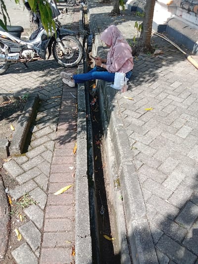 A young woman, wearing a pink hijab, sits on the edge of a concrete drainage ditch, using a cell phone. She is wearing blue jeans and pink and white shoes. Her left leg is extended, and her right leg is bent at the knee. She is positioned on a paved sidewalk, with the ditch running parallel to the sidewalk. There is a motorcycle parked behind her to the left. The ditch is surrounded by grey brick pavers with a herringbone pattern. The setting is a city street in Kendal, Indonesia, at the coordinates -6.931 latitude and 110.204 longitude. There is debris and dirt in the drainage ditch. The sidewalk is mostly shaded. The ground is covered with a layer of dried leaves.