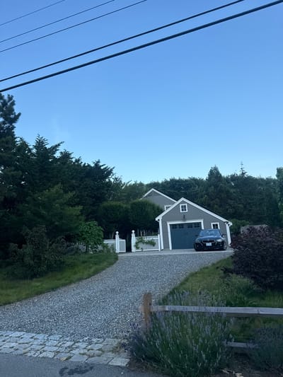 The image depicts a residential scene with a two-story house featuring a gray exterior and a dark gray garage door. A black BMW sedan is parked in the driveway in front of the garage. The driveway is made of gravel, and a white picket fence with decorative posts lines the property entrance. Lush green trees and bushes surround the house and driveway, creating a natural, wooded environment. In the foreground, a rustic wooden fence runs horizontally, with lavender plants visible in front of it. Overhead, power lines are strung across a clear blue sky. The scene appears to be in Provincetown, USA, suggesting a coastal or rural setting common to the area. There are no people or animals visible in the image.