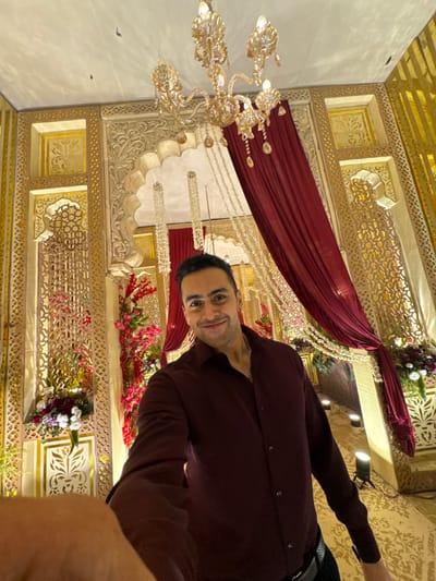 A man in a maroon shirt stands inside a decorated hall. The hall is adorned with ornate gold carvings and a maroon fabric hanging from the ceiling. There are chandeliers and flower arrangements as well. He smiles towards the camera.