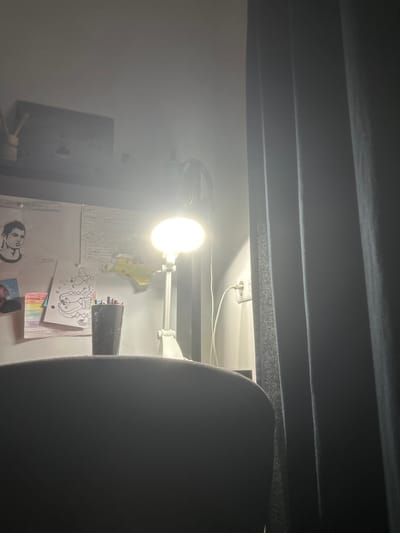 The image depicts a dimly lit desk area, likely in a bedroom or study. A bright desk lamp illuminates a section of the desk, revealing various items. On the desk, a black cup filled with colored pens and pencils is prominent. Several drawings and papers are pinned to a vertical surface behind the cup, including a sketch of a person's head, a rainbow-colored drawing, and a drawing of a dragon-like creature. A yellow cut-out of what appears to be Pikachu is also visible. To the right of the desk, a white electrical outlet with a white cord plugged in is mounted on the wall, partially obscured by dark grey curtains. The overall lighting suggests it is either evening or the room has limited natural light. The location is specified as Carrières-sous-Poissy, France.